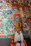 A statue of the Buddha, showing him as an emaciated renouncer relates to his enlightenment and his status as a yogic ascetic who has ultimate control over his body, Khinmey Nyingma Monastery, against a background of brightly coloured murals.